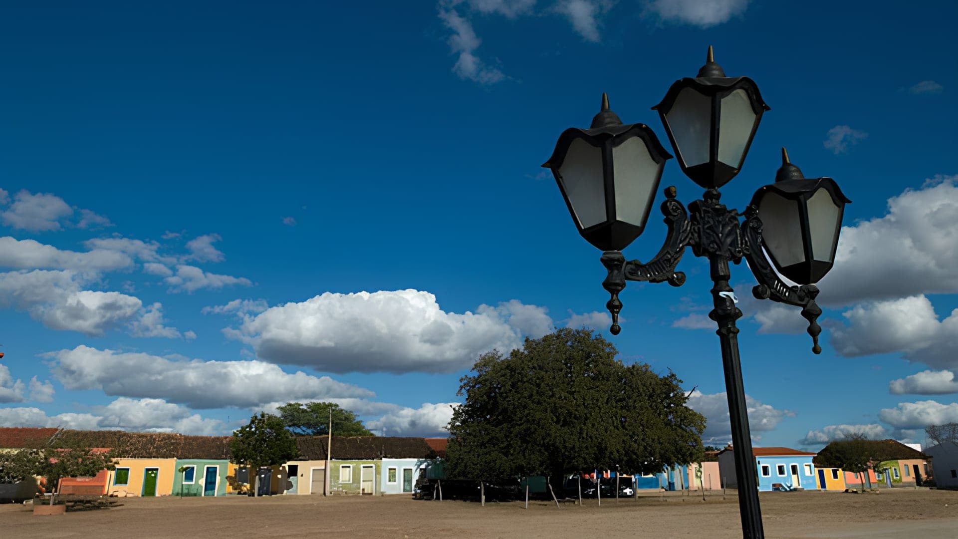 Tamarineiro centenário no largo de Caboclo sob céu azul, com um lampião de ferro em primeiro plano e os casarios ao fundo.